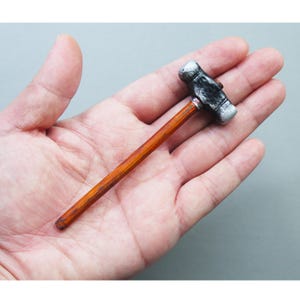 Könnte beinhalten: Ein Miniaturhammer mit silbernem Kopf und Holzgriff. Der Hammerkopf hat eine rechteckige Form mit dunkler, strukturierter Oberfläche. Der Griff ist ein schlanker, brauner Stab. Der Hammer wird in der Hand einer Person gehalten.