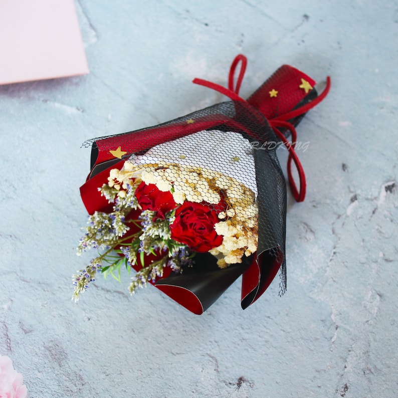 K&ouml;nnte beinhalten: Ein kleiner Strau&szlig; aus roten Rosen, Lavendel und Trockenblumen, verpackt in schwarzem Netz und rotem Papier. Der Strau&szlig; ist mit einem roten Band gebunden und hat goldene Sternakzente. Der Hintergrund ist eine hellblau-graue Oberfl&auml;che.