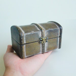 Peut inclure: Un petit coffre en bois de style antique avec un dessus incurvé et un fermoir en métal. Le coffre est brun vieilli avec des accents blancs, ce qui lui donne un aspect vintage. Le coffre est tenu dans une main.