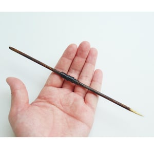 May include: A thin, brown wooden stick with a black band in the middle and a gold tip. The stick is held in a person's hand.