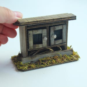 K&ouml;nnte beinhalten: Ein Miniatur-Holzstall mit zwei T&uuml;ren, jede mit einem Drahtgitterfenster und einem kleinen Knopf. Der Stall ist verwittert mit einer grau-braunen Oberfl&auml;che und steht auf einem Sockel, der mit gr&uuml;nem und braunem Moos bedeckt ist. Das Dach besteht aus kleinen Holzbrettern.