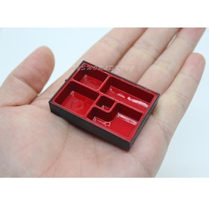 Peut inclure: Une petite boîte bento noire et rouge avec cinq compartiments. La boîte est en plastique et a une finition brillante.