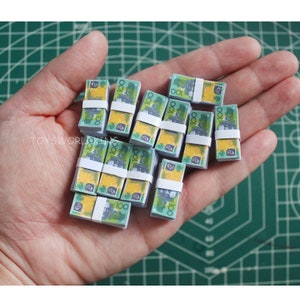 Op de afbeelding: Een hand die een stapel miniatuur Australische $ 100-biljetten vasthoudt. De biljetten zijn groen en geel met een portret van koningin Elizabeth II.
