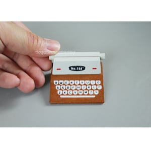 Puede incluir: Una máquina de escribir de madera en miniatura con la parte superior blanca y la base marrón. La máquina de escribir tiene una etiqueta roja que dice "No. 188'" y un teclado con teclas negras y letras blancas.
