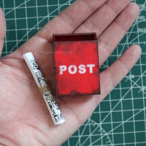 Puede incluir: Un buzón de madera rojo con la palabra "POST" impresa en blanco. Un pequeño trozo de papel enrollado está en la palma de una mano.
