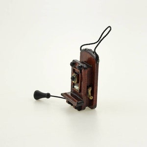 Pu&ograve; includere: Un telefono rotativo da parete in miniatura in legno con una cornetta nera e un filo nero. Il telefono ha una finitura marrone scuro e un gancio color ottone.