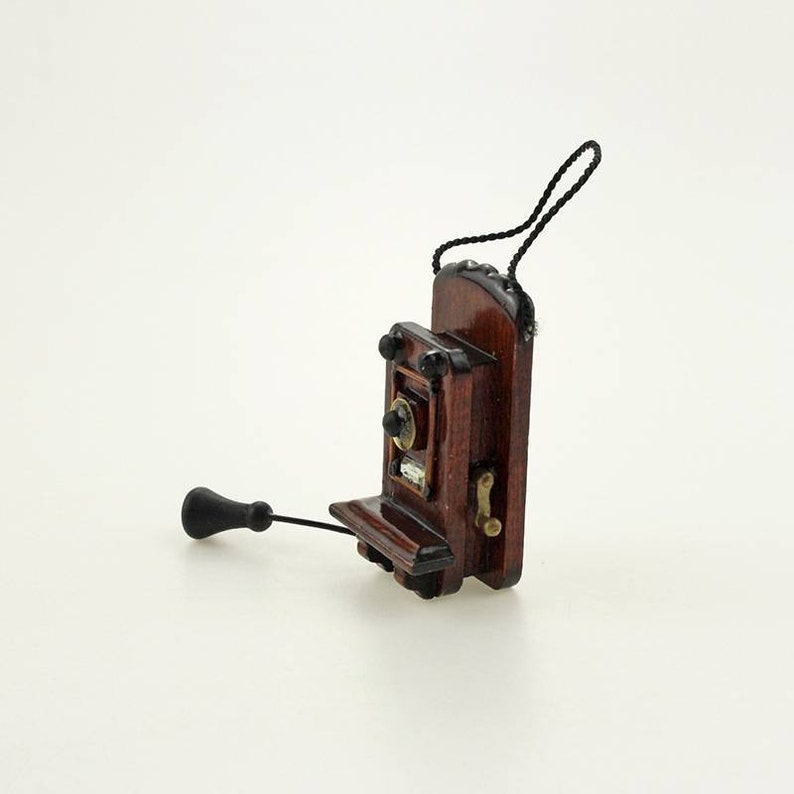 Pu&ograve; includere: Un telefono rotativo da parete in miniatura in legno con una cornetta nera e un filo nero. Il telefono ha una finitura marrone scuro e un gancio color ottone.