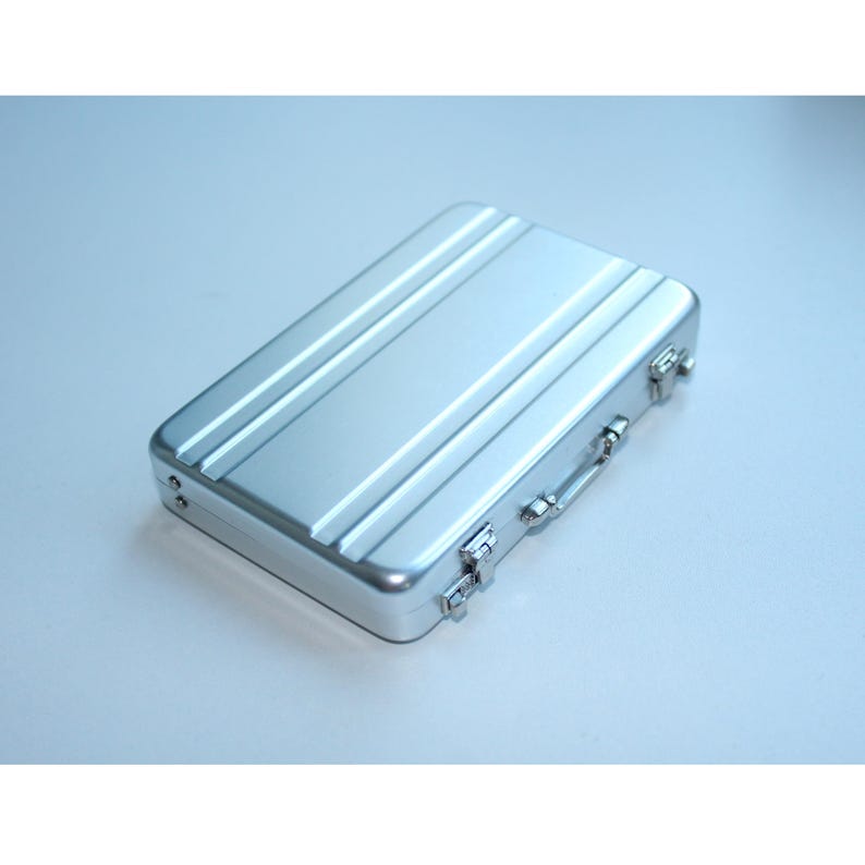 May include: A silver-colored, rectangular briefcase with a ridged top. The briefcase has a handle and two latches on the front. The case is made of metal and appears to be new.