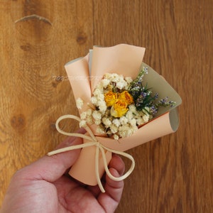 K&ouml;nnte beinhalten: Ein kleiner Strau&szlig; getrockneter Blumen, verpackt in pfirsichfarbenem Papier mit einem cremefarbenen Band. Der Strau&szlig; enth&auml;lt gelbe Rosen und wei&szlig;en Schleierkraut.