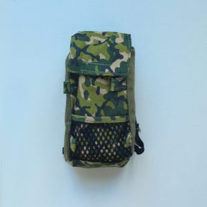 Puede incluir: Una pequeña mochila de camuflaje con un bolsillo de malla en la parte delantera. La mochila está hecha de una tela verde y marrón con un patrón de camuflaje.