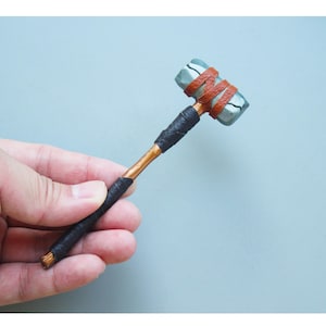Könnte beinhalten: Ein kleiner, handgefertigter Hammer mit einem Kupfergriff, der mit schwarzem und braunem Klebeband umwickelt ist. Der Hammerkopf ist aus grauem Ton und hat eine raue, strukturierte Oberfläche.