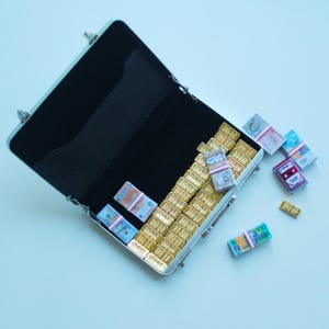 May include: An open silver briefcase with a black interior is filled with miniature gold bars and stacks of toy money. Several stacks of toy money and a few gold bars are scattered outside the briefcase on a light blue surface.
