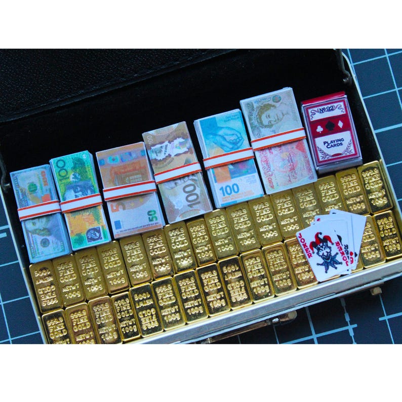 May include: A black briefcase filled with miniature gold bars, stacks of international currency, a deck of playing cards, and a joker card. The gold bars are labeled with "999.9 FINE GOLD". The currency includes US dollars and Euros.
