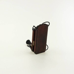 Pu&ograve; includere: Un telefono rotativo in miniatura di legno con un filo nero e una finitura marrone scuro. Il telefono &egrave; chiuso e la cornetta &egrave; appesa sul lato.