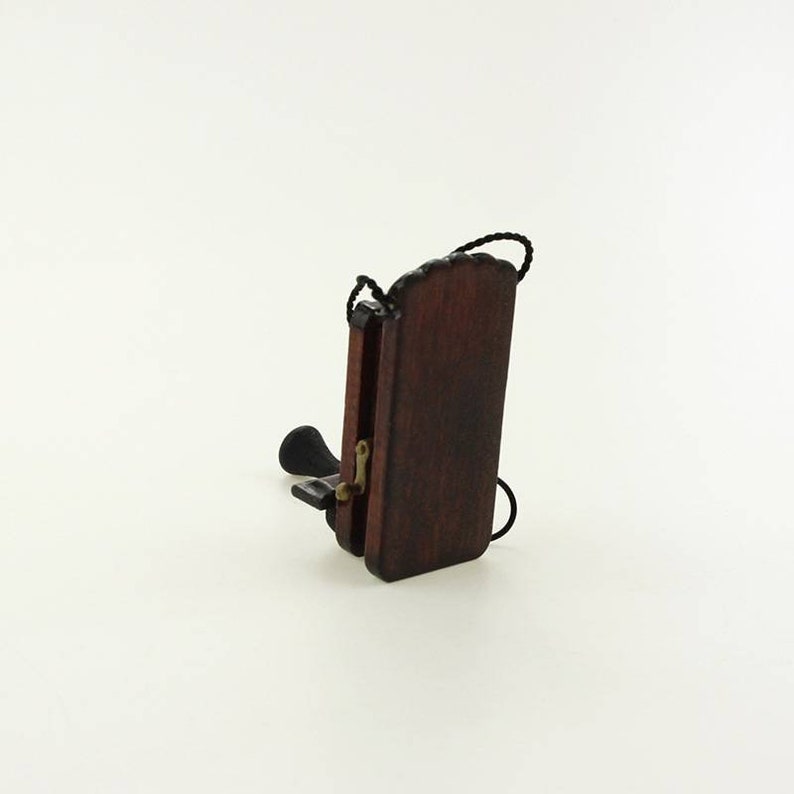 Pu&ograve; includere: Un telefono rotativo in miniatura di legno con un filo nero e una finitura marrone scuro. Il telefono &egrave; chiuso e la cornetta &egrave; appesa sul lato.