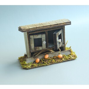 K&ouml;nnte beinhalten: Ein Miniatur-H&uuml;hnerstall aus Holz mit offener T&uuml;r, kleinem Fenster und schr&auml;gem Dach. Der Stall steht auf einer Basis aus gr&uuml;nem Moos und Stroh, mit drei orangefarbenen Eiern davor. Der Stall ist aus verwittertem Holz gefertigt.