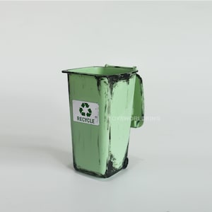 Può includere: Un bidone della spazzatura in metallo verde con un coperchio nero e un adesivo bianco che dice "RECYCLE" con un simbolo di riciclaggio verde. Il bidone ha un aspetto usurato con striature di vernice nera.