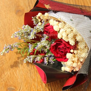 K&ouml;nnte beinhalten: Ein Strau&szlig; aus getrockneten Blumen mit leuchtend roten Rosen, zartem wei&szlig;en Schleierkraut und Lavendelzweigen mit lila Bl&uuml;ten. Die Anordnung ist in schwarzes und rotes Papier mit einem Netz &uuml;berzogen, was eine rustikale, aber elegante &Auml;sthetik schafft.
