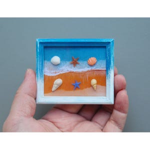 Könnte beinhalten: Eine Miniatur-Strandszene in einem quadratischen Rahmen. Das Kunstwerk zeigt einen blauen Himmel, einen sandigen Strand und kleine Muscheln und Seesterne. Der Rahmen ist blau und weiß bemalt und schafft ein Küstenmotiv.