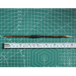 May include: A wooden dowel rod with a black and brown band in the center. The rod is about 10 inches long and is resting on a green cutting mat with a ruler underneath.