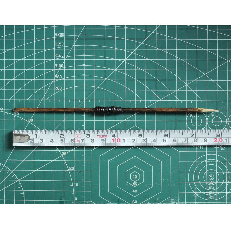 May include: A wooden dowel rod with a black and brown band in the center. The rod is about 10 inches long and is resting on a green cutting mat with a ruler underneath.