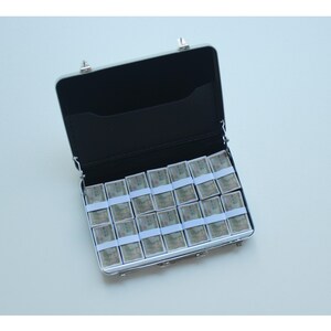 May include: An open, silver-colored briefcase filled with stacks of money. The stacks are wrapped in white paper and secured with white bands. The interior of the briefcase is black.