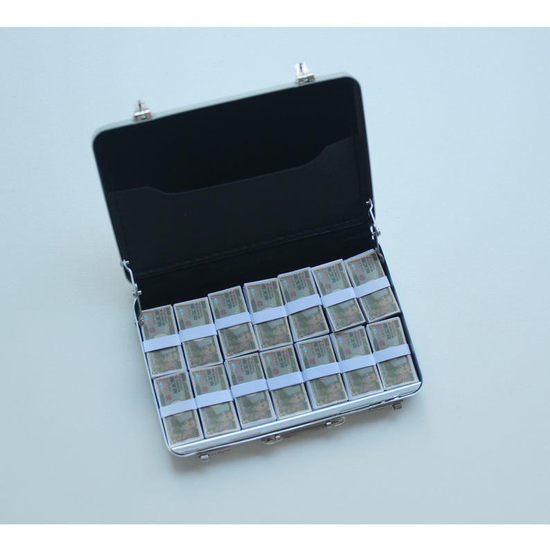 May include: An open, silver-colored briefcase filled with stacks of money. The stacks are wrapped in white paper and secured with white bands. The interior of the briefcase is black.