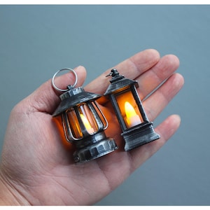 Peut inclure: Deux lanternes miniatures argentées avec des lumières LED jaune chaud. L'une a un anneau pour suspendre, l'autre est de forme classique. Les lanternes sont tenues dans une main sur un fond gris.