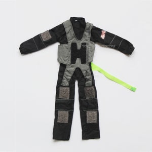 May include: Black and gray jumpsuit with a gray vest and a green strap. The vest has a black patch that says "GHOST FORCE".
