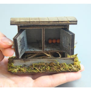 K&ouml;nnte beinhalten: Ein Miniatur-H&uuml;hnerstall aus Holz mit zwei F&auml;chern, jeweils mit einer Fl&uuml;gelt&uuml;r. Der Stall hat ein verwittertes Aussehen, mit einem moosigen Sockel und ein paar orangefarbenen Eiern im Inneren. Der Stall wird in einer Hand gehalten.