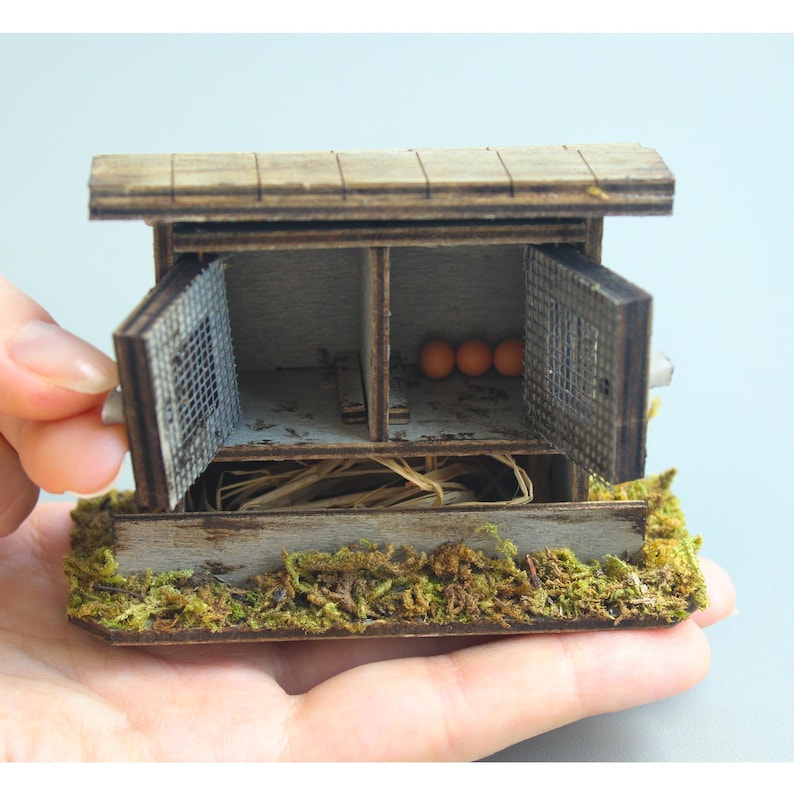 K&ouml;nnte beinhalten: Ein Miniatur-H&uuml;hnerstall aus Holz mit zwei F&auml;chern, jeweils mit einer Fl&uuml;gelt&uuml;r. Der Stall hat ein verwittertes Aussehen, mit einem moosigen Sockel und ein paar orangefarbenen Eiern im Inneren. Der Stall wird in einer Hand gehalten.