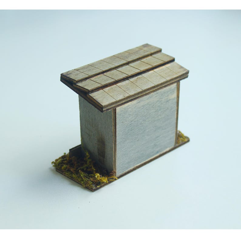 K&ouml;nnte beinhalten: Ein Miniatur-Holzgeb&auml;ude mit einer grauen Wand und einem braunen Schindeldach. Der Sockel ist mit gr&uuml;nem und braunem Moos bedeckt. Dieses kleine Geb&auml;ude ist wahrscheinlich ein Dekorationsartikel.