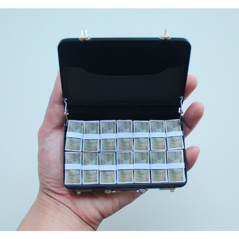 May include: A miniature black briefcase filled with stacks of tiny, rectangular items secured with light blue bands. The briefcase has silver clasps and hinges. The items inside appear to have a green and brown design.