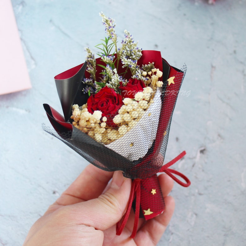 K&ouml;nnte beinhalten: Ein kleiner Strau&szlig; aus roten Rosen, Lavendel und wei&szlig;en Blumen, verpackt in schwarzem Netz und rotem Papier. Der Strau&szlig; ist mit einem roten Band gebunden. Das Geschenkpapier hat goldene Sterne.