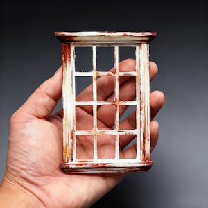 以下が含まれることがあります： 白と赤褐色の仕上げが施された、ミニチュアの風化した窓枠。窓には小さな正方形のガラスの格子があります。フレームは灰色の背景に対して手に持たれており、古びた素朴な美しさを表現しています。