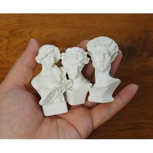 May include: Three white plaster busts of classical figures. The busts are on a wooden surface.