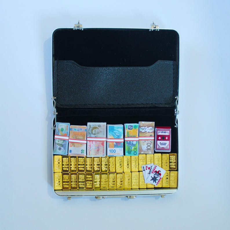May include: Open black briefcase filled with miniature gold bars, stacks of international currency, and a deck of playing cards. The briefcase is open, revealing a velvet interior.