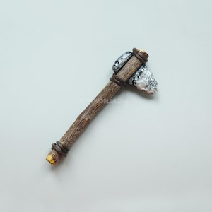 May include: A small, hand-crafted axe made from a wooden stick and a grey stone head. The axe is wrapped with brown wire at the handle and the head.
