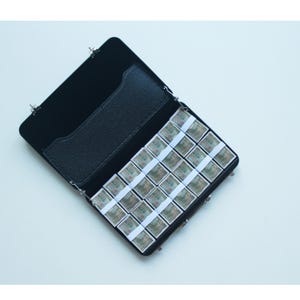 May include: Black leatherette case, open to reveal a grid of small, rectangular watercolor paint pans. The pans are arranged in rows and columns, each wrapped with a white paper band. The case has a black interior and a small pocket.