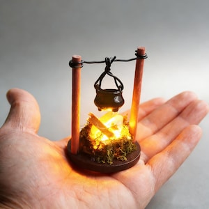 Könnte beinhalten: Miniatur-Lagerfeuerszene mit einem kleinen schwarzen Topf, der über einem glühenden Feuer hängt. Das Feuer besteht aus kleinen Holzstücken und Moos, das auf einer runden Holzbasis angeordnet ist. Zwei Holzpfosten stützen den Topf. Die Szene wird in einer Hand gehalten.
