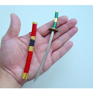 Könnte beinhalten: Zwei Miniaturschwerter, eines mit einem roten und goldenen Griff und das andere mit einem grünen und goldenen Griff, werden auf einer Hand gezeigt.