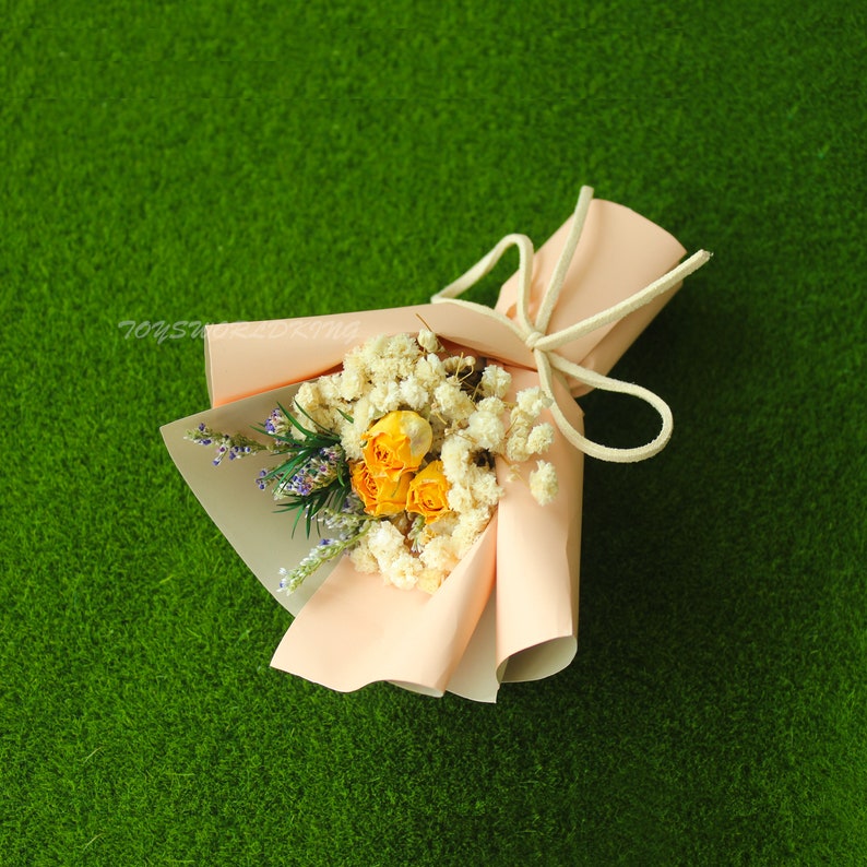 K&ouml;nnte beinhalten: Ein kleiner Strau&szlig; getrockneter Blumen, verpackt in hellrosa Papier mit einer wei&szlig;en Schleife aus Schnur. Der Strau&szlig; enth&auml;lt gelbe Rosen und wei&szlig;en Schleierkraut.