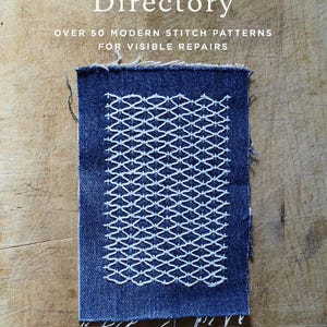 Könnte beinhalten: Das Buchcover mit dem Titel The Mending Directory: Over 50 Modern Stitch Patterns for Visible Repairs von Erin Eggenburg, herausgegeben von Search Press, zeigt ein rechteckiges Stück dunkelblauen Jeansstoff mit weißer geometrischer Stickerei.