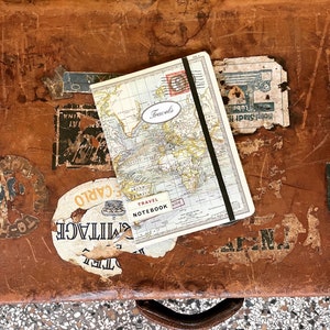 Könnte beinhalten: Ein vintage brauner Lederkoffer mit einem Reisetagebuch darauf. Das Notizbuch hat einen Weltkarten-Umschlag und die Worte "Reisetagebuch" und "Reisen" sind darauf gedruckt.