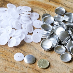 Puede incluir: Una colección de botones blancos de plástico y biseles de metal plateado se disponen sobre una superficie de madera. Los botones son redondos con un pequeño agujero en el centro. Los biseles metálicos también son redondos y tienen forma de cuenco.