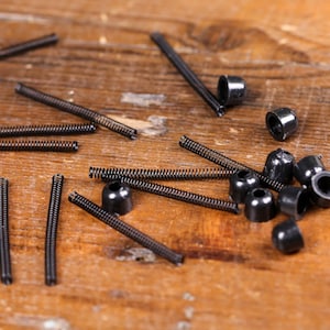 May include: A collection of small, black hardware components, including springs and cylindrical pieces, scattered on a weathered wooden surface. A coin is also present. The components appear to be for a mechanical device.