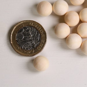 Könnte beinhalten: Eine Gruppe von 20 Naturholzkugeln mit einem Durchmesser von etwa 2,5 cm, die auf einer weißen Oberfläche verstreut sind. Eine britische Ein-Pfund-Münze dient als Größenvergleich.
