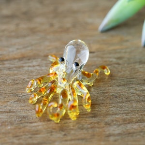 May include: A small glass octopus figurine with a clear body and brown spots. The octopus has eight tentacles and is sitting on a wooden surface.
