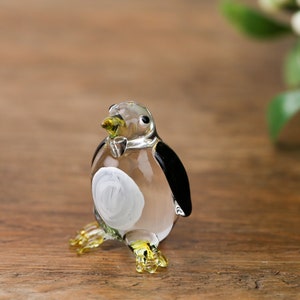 Può includere: Una piccola statuina di pinguino di vetro con la pancia bianca e le ali nere. Il pinguino è in piedi su una superficie di legno.