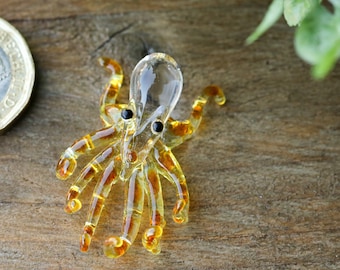 May include: A small glass octopus figurine with a clear body and amber-colored tentacles. The octopus has black eyes and is sitting on a wooden surface.